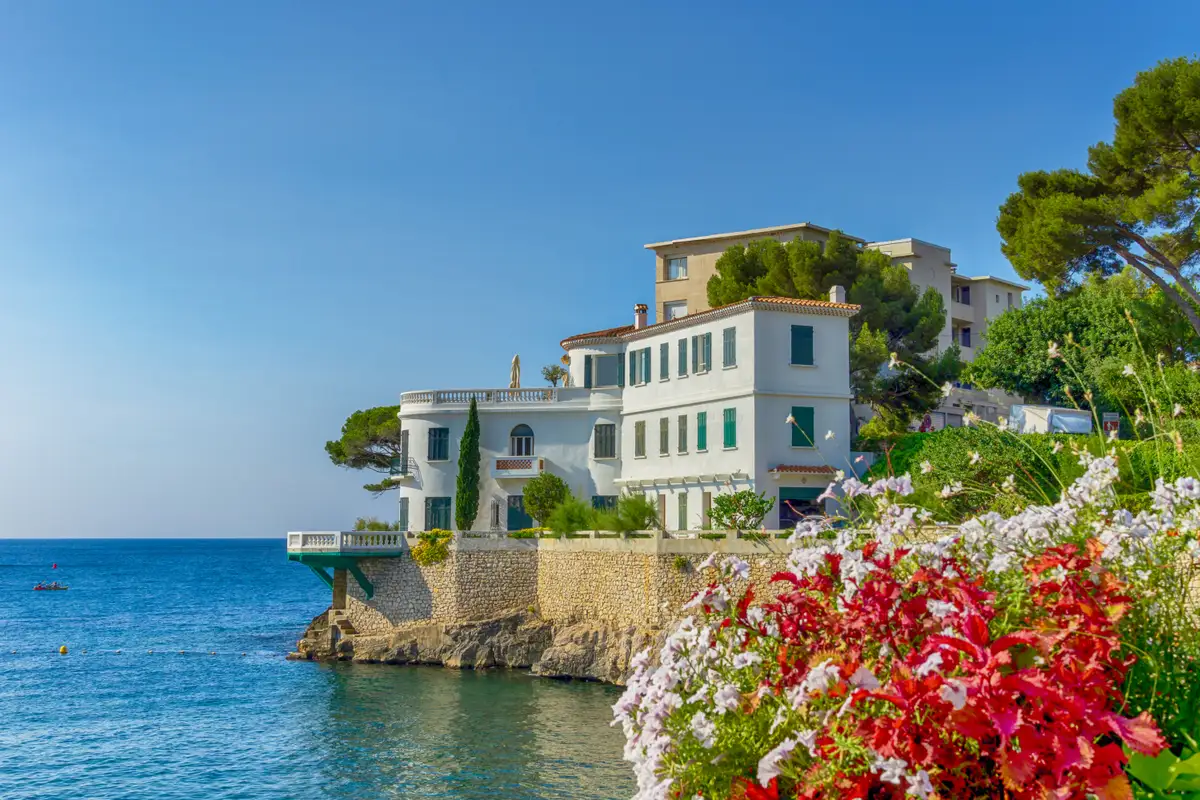Villa en bord de mer accrochée à la roche au-dessus d'une eau turquoise, massifs fleuris au premier plan